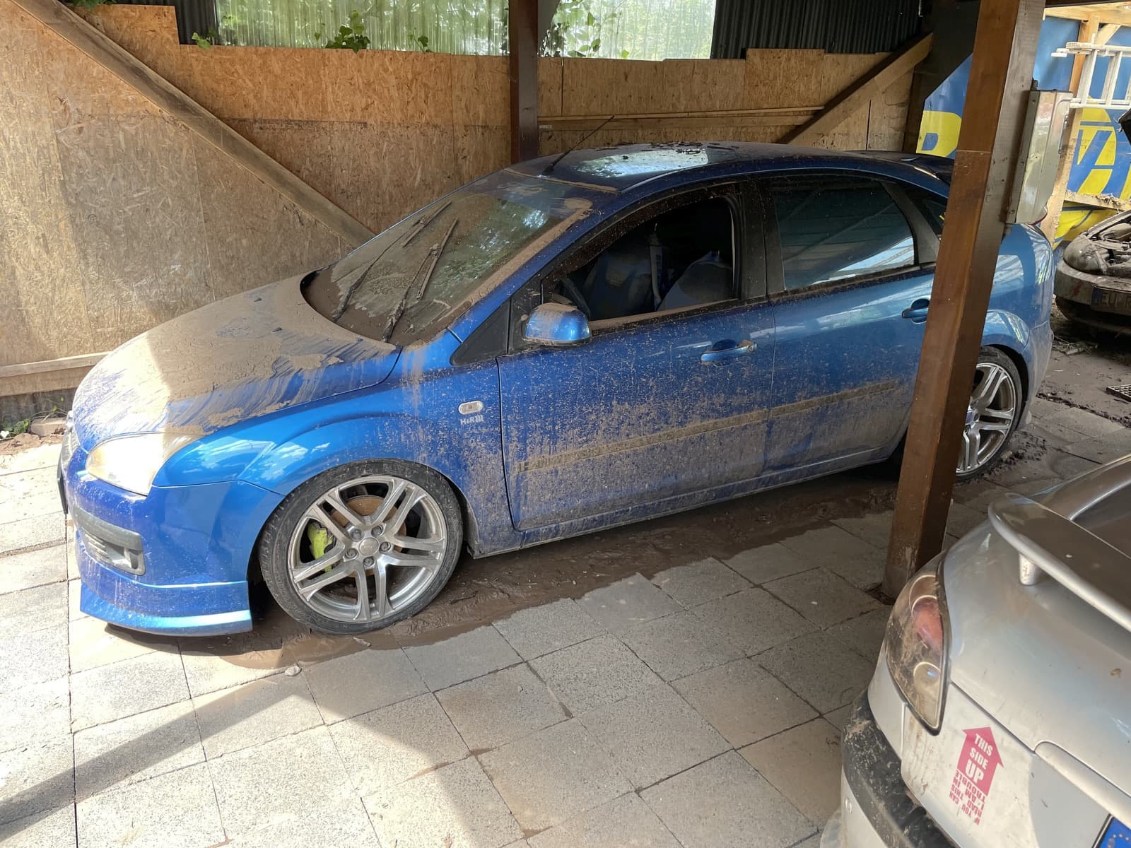 Von Schlamm überzogenes Auto unter einem Carport, daneben ein weiteres beschädigtes Fahrzeug