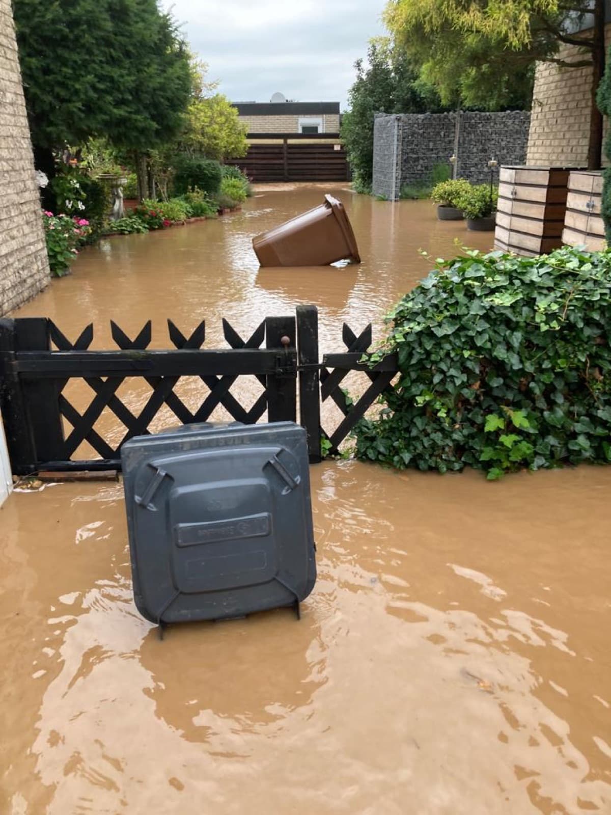 Überflutete Hofeinfahrt: braunes Wasser steht bis über das Gartentor, Abfalltonnen treiben darin