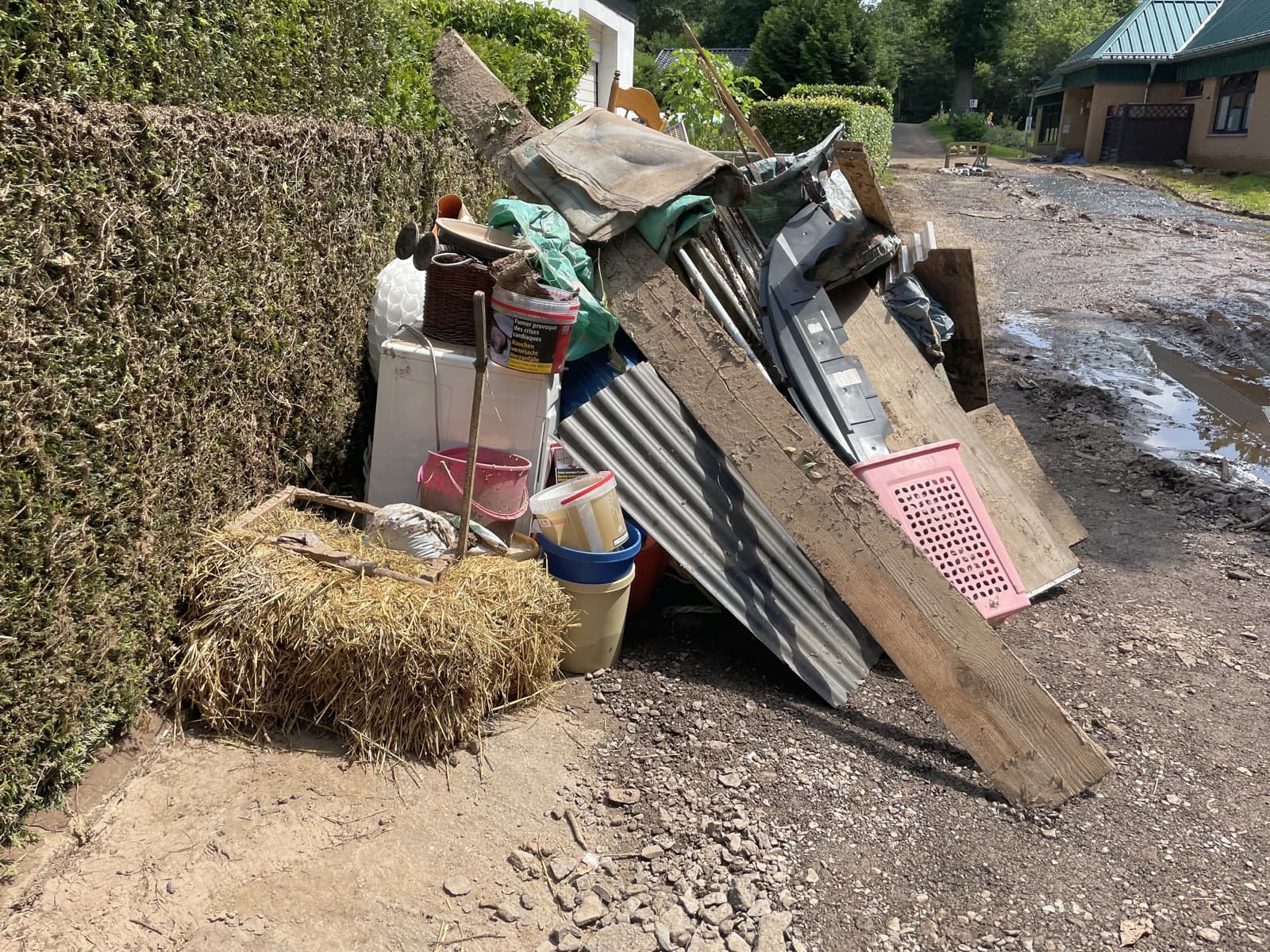 An einer Hecke aufgeschichtete Wellblechtafeln, Bretter und Eimer neben einer schlammigen Straße
